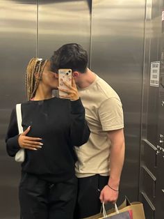 a man and woman kissing while holding shopping bags in front of a stainless steel elevator