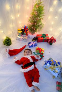 a baby dressed as santa clause laying on the floor next to presents and a christmas tree