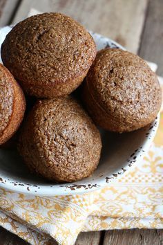 a white bowl filled with brown muffins on top of a wooden table next to a yellow napkin