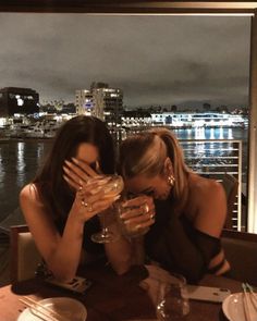 two women sitting at a table with wine glasses in front of their faces, looking out over the water