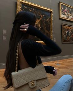 a woman sitting on the floor in front of paintings holding a beige purse and looking at her cell phone