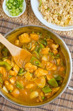 a pan filled with chicken and vegetable stew next to rice on a mat, along with a wooden spoon