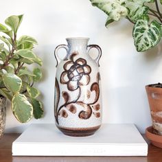 a white vase sitting on top of a wooden table next to potted plants and a book