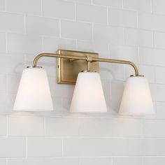 three light bathroom fixture with white glass shades on the bottom and gold finish, against a white brick wall