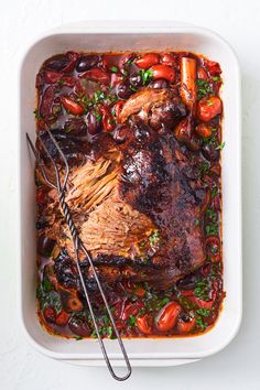 a roasting pan filled with meat, tomatoes and olives next to a pair of tongs