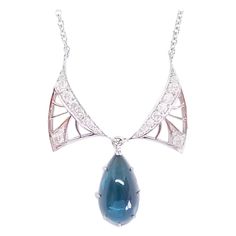 This Custom Designed and Crafted, One of a Kind Drop Necklace by Artisan Tom Castor features in Art Nouveau Tradition, Delicate and Open, Diamond Wings. Framed by the Wings is a Natural Indicolite Blue Tourmaline of 7.55 Carats in weight and a measurement of 10mm Wide x 18.5mm Long and a High Cabochon Depth of 5mm. The Gem is of AAA Quality and is held in an 18 Karat White Gold backed frame and prongs. The Drop and Wings are hinged for proper movement and placement on the Décolletage. The Diamon Art Deco White Gold Necklace With Gemstone, Art Deco White Gold Gemstone Necklace, White Gold Art Deco Gemstone Necklace, Diamond Cabochon Pendant Necklace, Art Deco Pendant Necklace For Formal Occasions, Luxury Pear-shaped Necklaces With Polished Finish, Luxury Pear-shaped Polished Finish Necklaces, Luxury Pear-shaped Necklace With Polished Finish, Luxury Pear-shaped Polished Finish Necklace