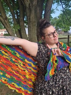 a woman wearing glasses is holding up a crocheted shawl in front of a tree