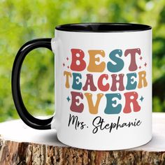 a black and white coffee mug with the words best teacher ever on it sitting on top of a tree stump