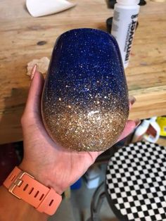 a hand holding up a blue and gold glittered wine glass on top of a wooden table