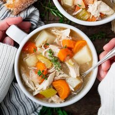 two hands holding bowls of chicken stew with carrots, celery and bread