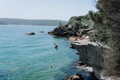 a person jumping into the water from a cliff near some rocks and trees, while others are swimming in the water