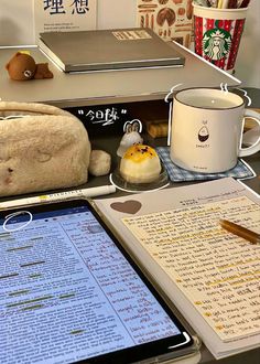 an open book on a desk next to a tablet and coffee cup with a pen in it