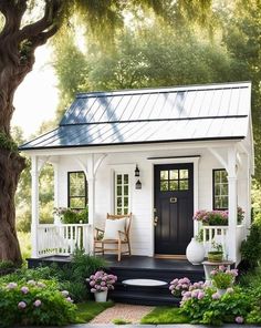 a small white house with black door and porch surrounded by greenery, flowers and trees