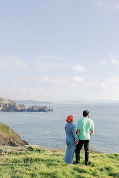 two people standing on top of a lush green hillside next to the ocean with an island in the background