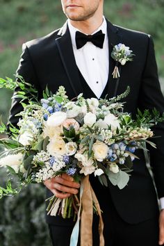 a man wearing a tuxedo holding a bouquet of flowers and greenery in his hand