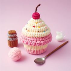 a crocheted cupcake with a cherry on top next to a spoon and ball of yarn