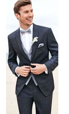 a man in a suit and bow tie standing on the beach with his hands in his pockets