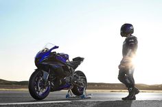 a man standing next to a blue motorcycle on the side of a road with sun shining behind him