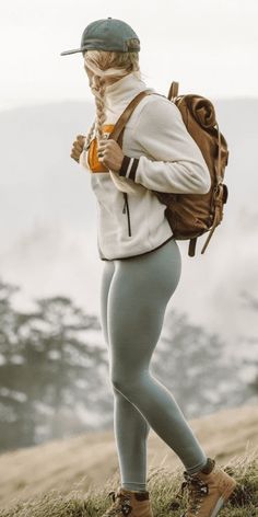 a woman standing on top of a grass covered hillside with a backpack and cell phone in her hand