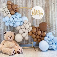 a teddy bear sitting in front of balloons and a happy birthday sign on a wall