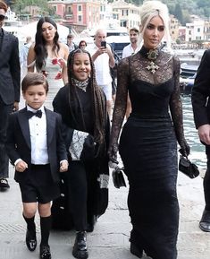 a woman in a black dress walking with two children and other people on the dock