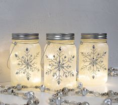 three mason jars with snowflakes on them are sitting next to silver bells and beads