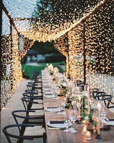 an outdoor dining area with long tables and lights strung from the ceiling over it, along with black chairs