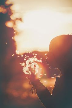 a woman holding a dandelion in front of her face as the sun sets