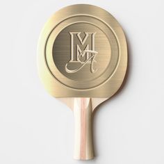 a gold ping pong paddle with the letter m on it's back side