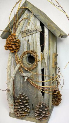 a birdhouse with pine cones and twine around it's neck, hanging on a white wall