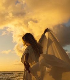 a woman in white dress standing on the beach at sunset with her arms spread out