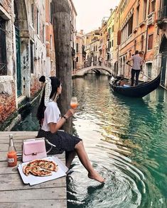 a woman sitting on a dock next to a river with pizza and drinks in front of her