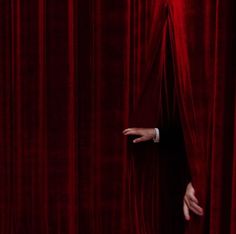 a man peeking out from behind a red curtain with his hand on the curtain edge