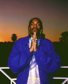 a man standing in front of a fence with his hands clasped to his chest while wearing a blue jacket