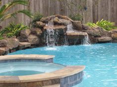 an outdoor pool with waterfall and rocks in the middle, next to a wooden fence