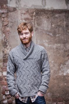 a man standing in front of a brick wall wearing a gray sweater and blue jeans