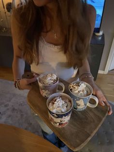 a woman holding two cups of hot chocolate and whipped cream on top of a wooden tray