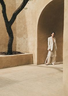 a woman in a white suit is standing near a tree and some concrete walls with an arch