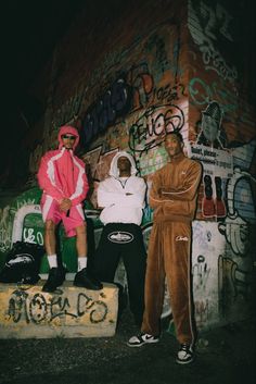 two men sitting on top of a cement block in front of graffiti covered wall with one man wearing a pink hoodie