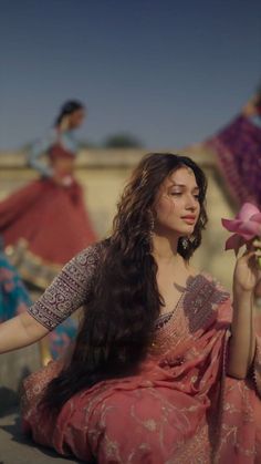 a woman sitting on the ground holding a pink flower in her hand with other women behind her