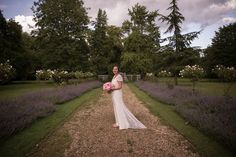 a woman in a wedding dress standing on a dirt road with lavender bushes and trees behind her