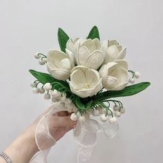 a hand holding a white flower bouquet with green leaves and flowers on it's side