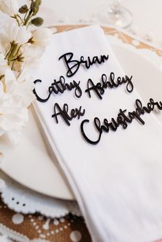 a white plate topped with a napkin covered in black writing next to flowers and wine glasses