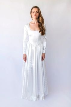 a woman wearing a long white dress and standing in front of a white wall with her hands on her hips