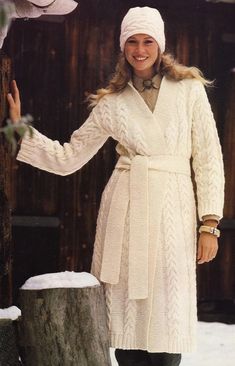 a woman wearing a white coat and hat standing next to a tree stump in the snow