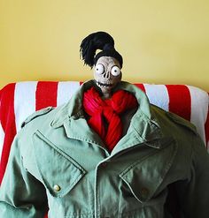 a creepy doll in a green jacket sitting on a red and white striped couch next to a yellow wall