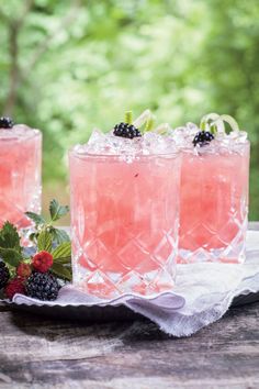 three glasses filled with ice and berries on top of a wooden table in front of trees