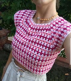 a woman wearing a pink and white crochet top standing in front of a bush