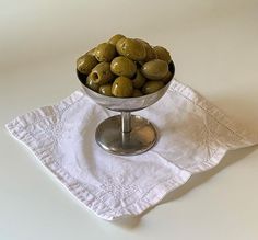 a glass bowl filled with green olives on top of a white cloth covered napkin