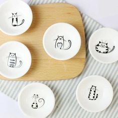 four white bowls with cats drawn on them sitting on top of a wooden tray next to a blue and white striped napkin